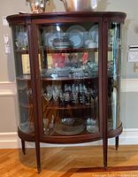 Front view of curved glass china curio cabinet showing curved glass panels, front locking door, wood frame, and displayed glassware and china dishes inside.