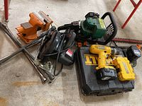 Overview photo showing all items together on floor: DeWalt drill, circular saw, hedge trimmer, vise, clamps, and lug wrench