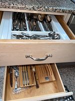 Two open drawers revealing assorted stainless steel flatware and serving utensils, with some smaller knives and forks in a wooden rack organizer.