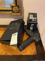 Photo showing GoPro Hero 3 camera with waterproof housing in original box placed behind the Hypervolt Plus handheld massager on a wooden table.