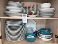 Two shelves showing multiple stacks of white plates, bowls, and serving ware along with some glassware and lidded containers. Items are ceramic and glass, mostly white with some teal and floral accents.