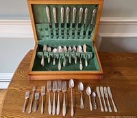 Opened wooden box with green velvet interior holding a set of silver plated flatware including dinner knives, forks, and spoons. Additional flatware is spread on table in front of the box.