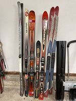 Full view of all the skis standing upright including cross country and downhill skis with varying lengths, plus a black hard case on the side