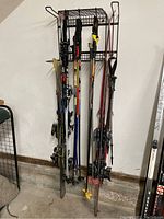 Photo of skis and ski poles arranged on a metal rack against a wall, showing the overall quantity and condition of items.