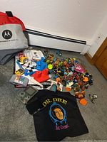 Wide shot showing a Motorola branded backpack, assorted small toys and figurines, two Nintendo handheld consoles, and a black Dr. Dre graphic T-shirt.