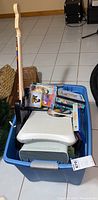 Blue storage bin filled with a white Xbox 360 console, Wii Fit board, video game discs and cases, and a Guitar Hero guitar controller with multicolored fret buttons.
