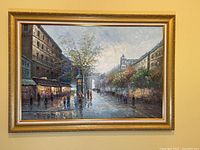 Full view of the framed street scene painting hung on a wall, showing the entire artwork and its gold frame.