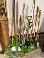 Wide shot showing assortment of yard tools including shovels, picks, pitchfork, leaf rakes with green heads and wooden handles, and other tools leaning against wall.