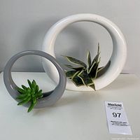 Two round ceramic vases, one large white and one smaller gray, each with faux plants inserted, shown from a front angle on a gray surface against a gray wall.