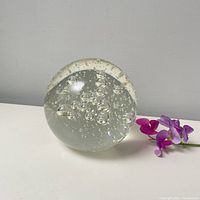 Full view of the round clear glass paperweight with visible controlled bubbles inside, placed on a white surface beside a purple flower.