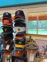 Two tall stacks of multiple sports caps arranged on a rack. Caps feature various logos from baseball and football teams including New York Yankees, Boston Red Sox, USC, Pittsburgh Pirates, Oakland Athletics, Texas Longhorns, and more.