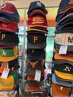 Rack of sports caps showing various team logos and colors stacked vertically on a rack in front of a window.