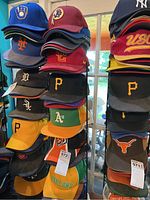 Rack showing stacks of sports caps with logos from baseball and football teams including Brewers, Mets, Tigers, White Sox, Pirates, Athletics, Redskins, and others.