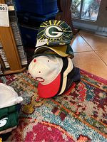 Stack of multiple sports caps on a patterned carpet including a Green Bay Packers cap with large embroidered 'G' logo and yellow sunray design