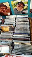 Wooden rack filled with rows of vertically standing music CDs, showing various album spines and titles, with a background featuring two portraits of men.