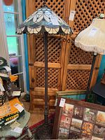 Full view of Tiffany style floor lamp with metal base and multicolored stained glass shade, standing next to wooden cabinet and other lamps.
