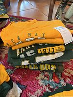 Three folded Green Bay Packers T-shirts in yellow and green with vintage and player graphics stacked on a colorful rug.
