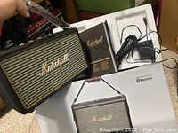 Marshall Bluetooth portable speaker held by a hand alongside its original box, power adapter, and instruction manual