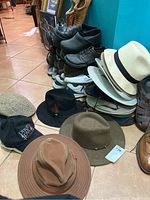 Various men's hats stacked and placed on floor near a shoe rack. Includes fedoras, outback hats, and caps in assorted neutral colors.