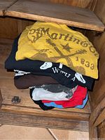 Stack of mixed music-themed printed T-shirts folded inside a wooden cabinet, showing the variety and condition of the lot.