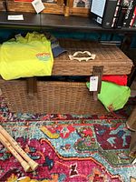 Photo showing wicker trunk with leather straps and scratches at the corner. Several folded Ron Jon T-shirts in yellow, green, red visible inside and on top of the trunk.
