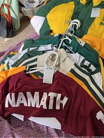 Photo showing several sports jerseys and sweatshirts including a maroon Namath jersey and a yellow and green numbered jersey with other green and cream apparel underneath.