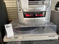 Photo showing the Technics turntable system under a cassette/dvd player in a silver casing, entire dust-covered dust cover and front edge turntable base visible.