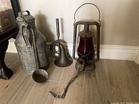 View of the three items together showing the milk jug with ladle, brass horn, and Dietz lantern with red glass globe and chain lock