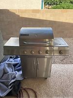 Full front view of stainless steel Perfect Flame barbecue grill with four control knobs, double-door storage underneath, and side shelves. A folded cover and hose are seen beside the grill.
