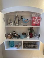 Photo of white cabinet shelves with assorted glass and ceramic holiday-themed decorative items including snowmen and bird figurines, music boxes, trinkets, small gingerbread house, and tissue box cover.
