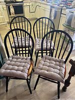 Four wooden chairs with black spindle backs and natural wood seats with beige tufted cushions tied on. Minor scuffs visible on chairs.