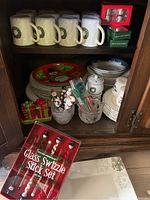 Shelf with white Mikasa Santa face cups, Christmas themed plates, salt and pepper shakers shaped like gifts, snowman swizzle sticks in glass, and Christmas themed spreaders