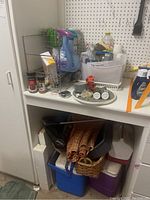 Overview photo showing various cleaning bottles, basket, folded rugs, Igloo coolers, and DustBuster under a desk.