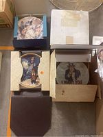 Box of assorted collector plates showing some plates in packaging, including an Uncle Sam plate and another round plate in blue and white box.