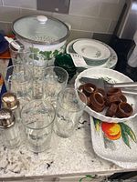 Full view of lot contents showing Corelle ivy print plates in two sizes, double boiler pot, six ivy printed drinking glasses, wooden napkin rings, pie servers, peach print platter, rose print pie dish, and plastic dessert cups on a kitchen counter.