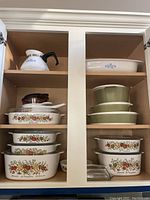 Photo of white Corningware ceramic cookware with vegetable pattern and lids, green ceramic bakeware, and white Pyrex dish inside kitchen cabinet