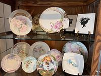 Multiple decorative porcelain plates displayed on shelves, mostly round with floral designs and one square plate with blue floral high heel shoe. Includes two black silhouette portraits displayed on small easels.