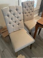 Two beige tufted cloth dining chairs side by side showing front and back tufted design with wooden legs.