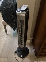 Silver and black tower fan with circular base, standing on wooden floor next to furniture.