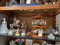 Display cabinet shelf showing porcelain woman figurine, musical carousel horses, trinket dishes, small decorative containers, and crystal carousel dish.