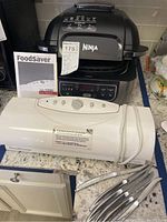 Photo showing Ninja Foodi Grill with user manual, FoodSaver Vac1200 vacuum sealer with user manual, and six knives on countertop.