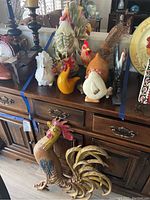 Rooster decorations on wooden cabinet including ceramic, handmade stick and hay, blown glass, plastic wall art, wooden carved roosters and small nest with eggs.