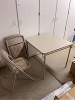 Photo of the folding table and set of four chairs. The table is square with a vinyl tabletop and metal legs. The chairs are folded and stacked beside the table.