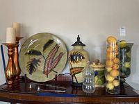 Overview of lot showing decorative ceramic vases, stackable glass containers filled with faux lemons and limes, decorative plate with leaf design, and candlestick holders with candles