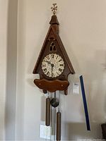 Full view of the vintage wooden cuckoo clock hanging on a white wall showing the pointed roof, clock face with Roman numerals, brass weights, and pendulum.