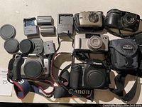 Top down view of all cameras including four Canon cameras and one Olympus camera, with accessories such as battery chargers and lens caps.