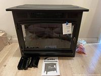 Front view of black Duraflame electric fireplace with glass front, user manual and accessory trays placed in front.