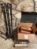 Set of four black metal fireplace tools standing on stone floor next to a stone fireplace, with an open cardboard box containing six Duraflame fire logs visible.