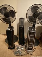 Front view of 2 Lasko fans, 2 Lasko heaters, 1 Aikoper heater, and 5 Lasko remotes in bag on carpeted floor