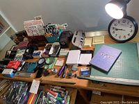 Overview photo showing a large collection of office supplies and stationery on a wooden desk including organizers, writing tools, cutting mat, clock, lamp and greeting cards.